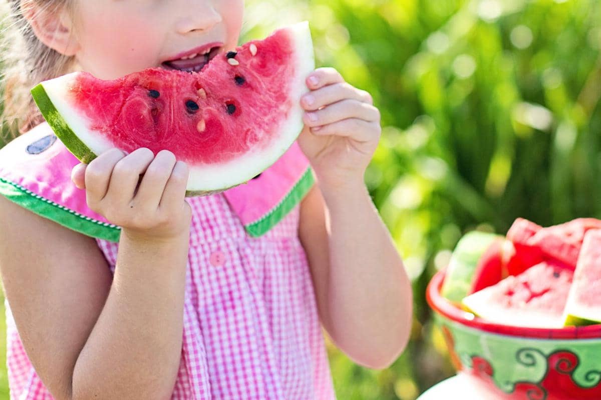 Anak kecil mengenakan gaun merah muda sedang memakan sepotong besar buah semangka segar di luar ruangan pada hari cerah.